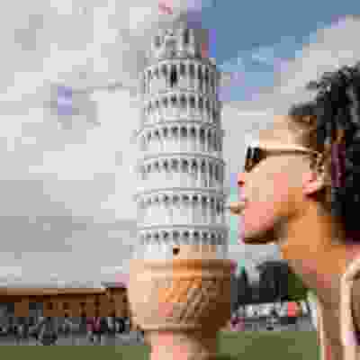 Women sticking her tongue out at the leaning tower of pisa, holding an ice-cream cone beneath it.